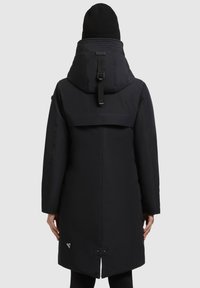 Fekete vízálló parka kapucnival, magas gallérral, állítható pántokkal és logó részlettel az alján. Simább textúrájú, térd hosszúságú.