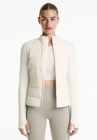 Femme aux cheveux attachés portant une veste rembourrée courte crème, un haut à manches longues et des leggings gris avec des rayures latérales sur fond blanc.