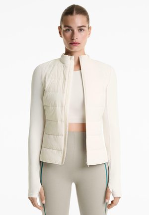Donna con i capelli tirati indietro che indossa una giacca imbottita beige corta, una maglietta a maniche lunghe e leggings grigi con strisce laterali su uno sfondo bianco.