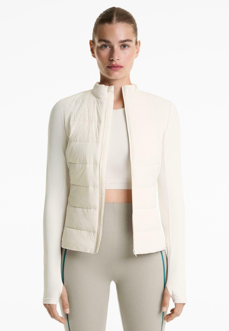 Femme aux cheveux attachés portant une veste rembourrée courte crème, un haut à manches longues et des leggings gris avec des rayures latérales sur fond blanc.