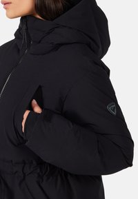 Kvinde iført sort polstret jakke med hætte, der justerer brystlommen, viser tommelfingerhul på ærmekant og logo-patch på armen.