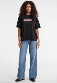 Žena s kovrčavom kosom koja nosi crnu majicu s ružičastim logotipom "Guess Jeans", plave široke traperice i crne cipele, stoji nasuprot bijeloj pozadini.
