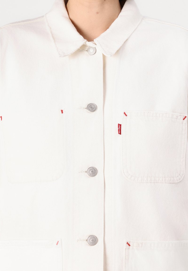 Veste en denim blanc avec un col côtelé, boutons-pression argentés, poches avant avec des accents de surpiqûres rouges et texture douce.