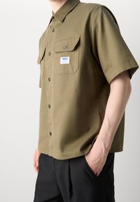 Olivegrünes Kurzarmhemd aus einem Baumwollmix, mit zwei Brusttaschen, Knopfleiste und einem Etikett auf der linken Tasche.