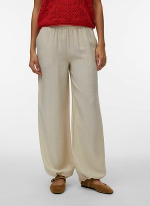 Moteris vilki laisvus smėlio spalvos kelnes su surinktomis čiurnomis, derančias su rudais lygiais batais ir rankovių neturinčiu raudonu megztu viršutiniu drabužiu.