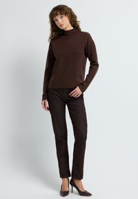 Vrouw met krullend haar in een bruine coltrui, bijpassende donkerbruine broek en bruine hoge hakken, staande tegen een effen achtergrond.