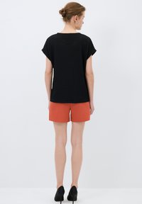 Top nero texturizzato con maniche corte, abbinato a pantaloni arancioni. La modella indossa tacchi neri, in piedi su uno sfondo bianco uniforme.
