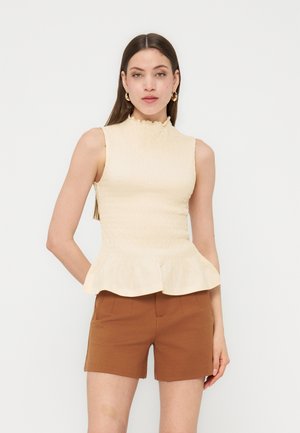 Vrouw die een mouwloze crèmekleurige blouse met gerimpelde zoom en bruine, op maat gemaakte korte broek draagt, staand tegen een effen lichte achtergrond.