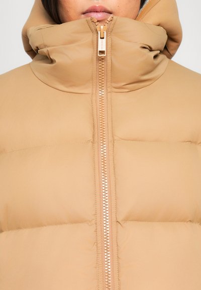 Nahaufnahme einer Person, die eine beigefarbene gepolsterte Jacke trägt, die bis zum Kinn zugezippt ist, mit einem hohen Kragen, der den Hals bedeckt.