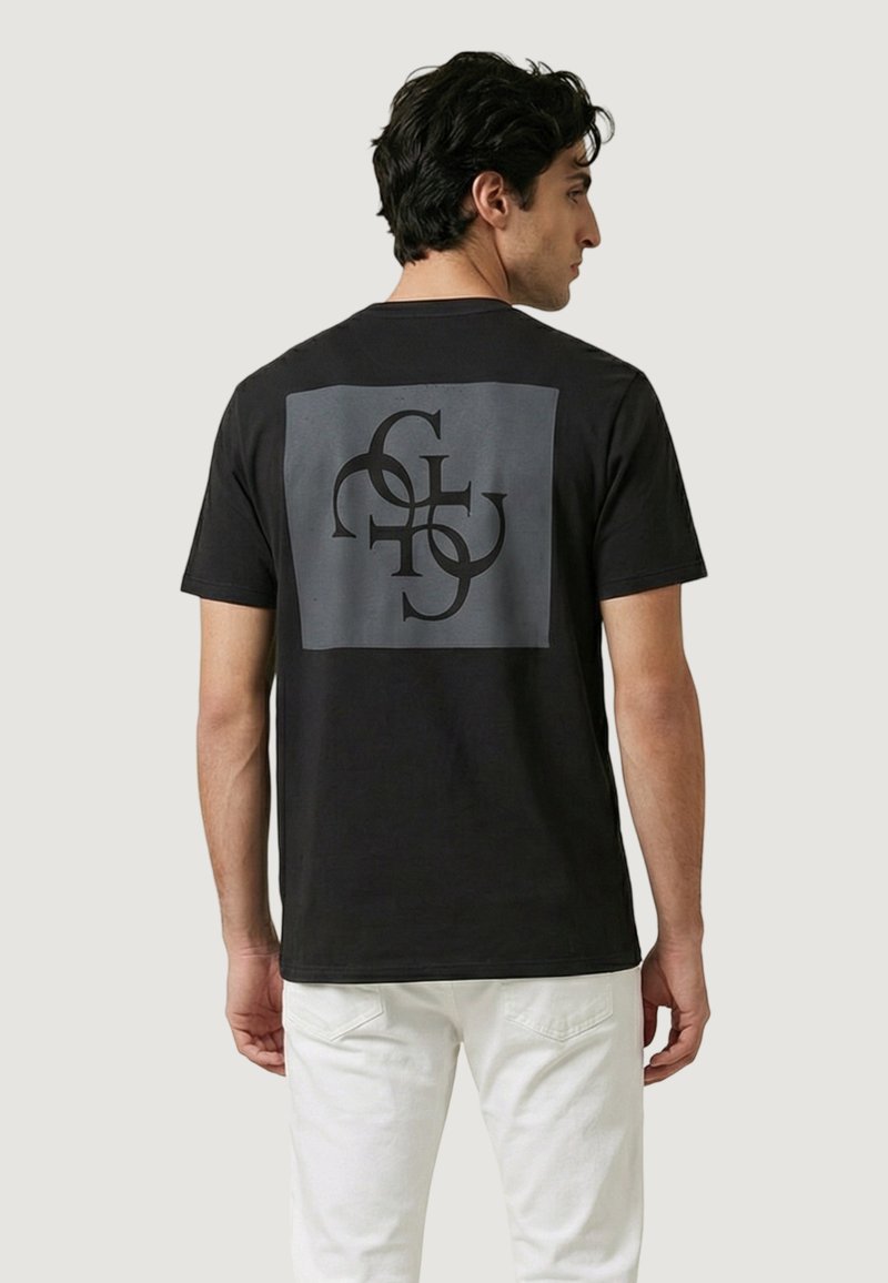 Hombre con camiseta negra con letras negras abstractas en un cuadrado gris en la parte trasera, combinado con pantalones blancos, de pie contra un fondo neutro.