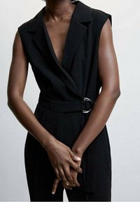 Black sleeveless jumpsuit with a deep V-neck and a wrap design, featuring a silver ring detail on the waist and smooth, tailored fabric.
