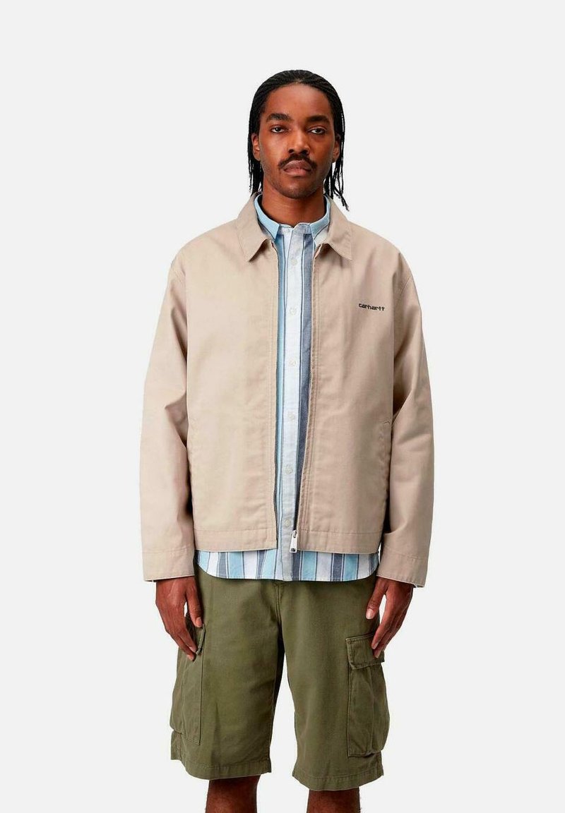 Chaqueta beige clara con cremallera y dos bolsillos laterales, llevada sobre una camisa de rayas azul claro y blanca y pantalones cortos de cargo en color verde olivo.