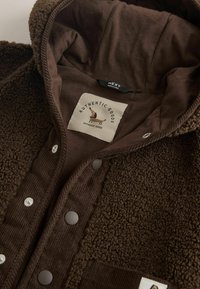 Brun texturerad fleecejacka med huva, tryckknappar och etikett med en liten hund och texten "AUTHENTIC GOODS."