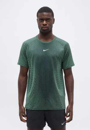 Mann steht, trägt grünes gemustertes Nike Sportshirt und schwarze Shorts mit Nike-Logos, neutraler Gesichtsausdruck, vor einfachem hellem Hintergrund.