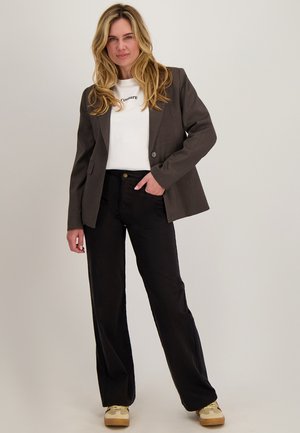 Vrouw met een donkerblazer, wit bedrukt shirt, zwarte broek en beige sneakers, staand met één hand in de zak tegen een effen achtergrond.