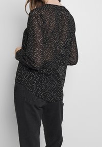 Blusa nera trasparente con pois bianchi, maniche lunghe, scollo rotondo e orlo curvo. Il tessuto è leggero e leggermente traslucido.