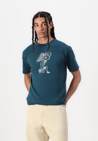 Kortærmet teal bomulds T-shirt med et hvidt grafisk design, der viser en omridsfigur og bogstavet "B" i bevægelse. Båret med lyse bukser.