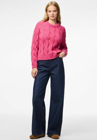 Vrouw staat met één hand in de zak, draagt een roze kabelbrei trui, donkere wijde jeans en bruine schoenen tegen een witte achtergrond.