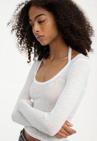 Top bianco a maniche lunghe realizzato in un tessuto leggero e semitrasparente, con scollo a barca e design aderente, indossato con le braccia incrociate.