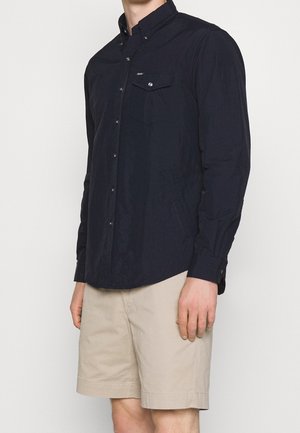 Mand iført sort langærmet skjorte med brystlomme og beige knælange shorts, stående mod en ensfarvet hvid baggrund.