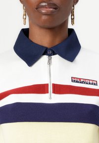 Hvid zip-polo med marinefarvet krave og røde, marinefarvede og gule vandrette striber. Har et "Hilfiger Sailing Gear" logo på brystet.