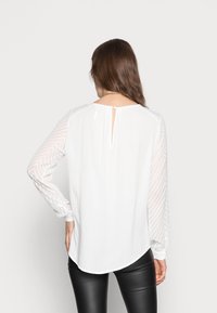 Blouse blanche avec des manches transparentes à motif en chevrons, un ourlet arrondi et un détail en forme de trou de serrure au dos. Le tissu semble léger et lisse.