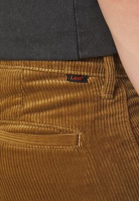 Gros plan sur un pantalon en velours côtelé brun avec l'étiquette de la marque Lee cousue au-dessus d'une poche arrière, porté avec une chemise gris foncé.