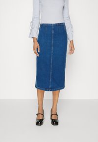 Faldă tip creion din denim albastru, cu croială dreaptă, având o cusătură frontală și un tiv care atinge jumătatea gambei; asortată cu un top cu mâneci lungi de culoare albastru deschis.