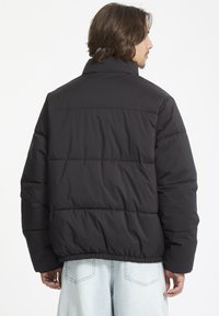 Zwarte pufferjas met hoge kraag, gewatteerd ontwerp, elastische zoom en lange mouwen, weergegeven vanaf de achterkant tegen een witte achtergrond.