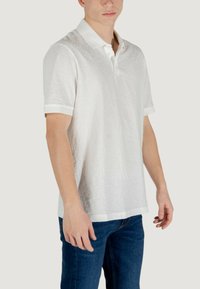 Polo shirt bianco a maniche corte con un motivo testurizzato, colletto classico e polsini bianchi a contrasto, abbinato a jeans blu scuro.