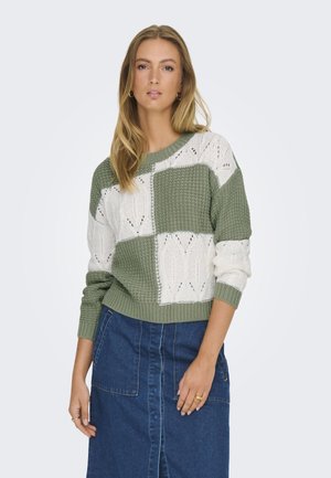 Strickpullover mit Patchwork-Design in Grün und Weiß, aus strukturiertem Stoff und mit lockeren Ärmeln. Getragen über einem Jeansrock.