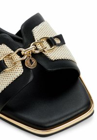 Black slide sandal with a woven cream and beige upper, gold chain accent, and a square toe. Features a metallic charm detail.