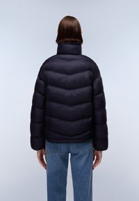 Giubbotto imbottito navy con imbottitura a chevron, colletto alto e polsini a costine. Indossato sopra jeans azzurri chiari. Texture del tessuto liscia e lucida.