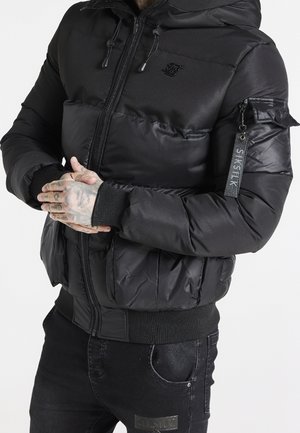 Mand iført sort polstret jakke med hætte og lynlås, mærkeetiket på ærmet, hænderne foldet, viste tatoveringer, iført sorte jeans.