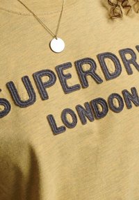 Gul sweatshirt med grå upphöjd text "SUPERDRY LONDON" och ett silverfärgat runt hängsmycke på en persons bröst.