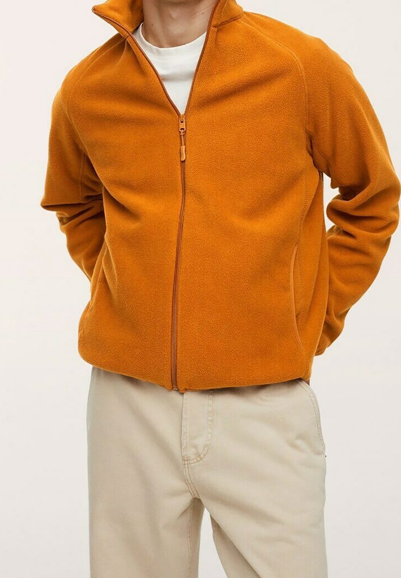 Homme portant une veste polaire orange zippée par-dessus une chemise blanche avec un pantalon beige, les mains dans le dos, debout devant un fond uni.