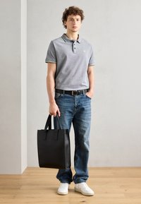 Camisa polo gris con acentos en negro y amarillo, jeans azules, zapatillas blancas y una bolsa tote negra con asas planas. De pie sobre un suelo de madera.