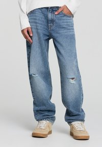 Jeans in jeans chiaro, di denim usurato, con una vestibilità ampia, caratterizzati da strappi all'altezza del ginocchio. Abbinati a sneaker beige e a un top bianco a maniche lunghe.