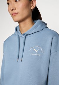 Lys blå hettegenser laget av myk tekstil, med kengurulomme og brodert "PUMA ATHLETICS"-logo med en hoppende puma.