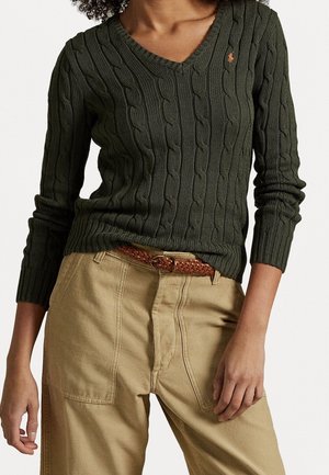 Mujer con un suéter de punto grueso color verde oliva con cuello en V y pantalones beige, con un cinturón marrón tejido, de pie con una mano cerca de la cadera.