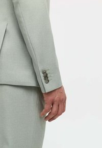 Hellgraue Anzugjacke mit strukturiertem Muster, ausgestattet mit vier kleinen Knöpfen am Ärmel und scharfer Schnittführung. Sichtbare Hand, die auf dem Ärmel ruht.