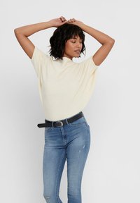 Cream ribbed short-sleeve top paired with light blue jeans and a black belt. Model poses with arms raised against a plain background.