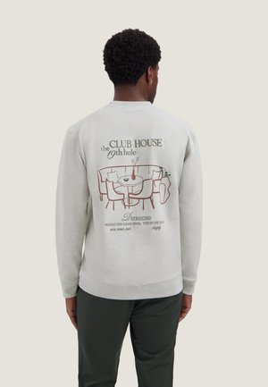 Man draagt een lichtgrijze sweatshirt met een illustratie van een clubhuis en tekst op de rug, gecombineerd met een donkere broek, met de rug naar de camera toe.