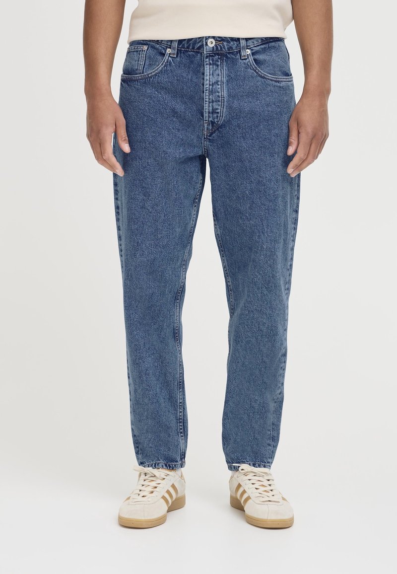 Personne portant un jean bleu droit et des baskets blanches avec des rayures beige, debout devant un fond clair uni.