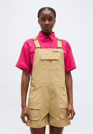 Γυναίκα που φοράει κοντό ταμπά σαλόπετα με μπροστινές τσέπες πάνω από φωτεινό ροζ μπλουζάκι με κοντά μανίκια και γιακά, στέκεται μπροστά σε απλό ανοιχτό φόντο.