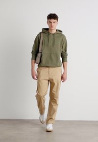 Felpa verde oliva con tasca frontale, abbinata a pantaloni beige e sneaker bianche. Il modello porta una borsa a tracolla grigia.