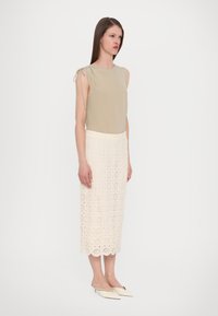 Haut sans manches beige avec des liens aux épaules, associé à une jupe blanche en dentelle arrivant au genou avec des motifs en découpe circulaire et un ourlet en vagues.