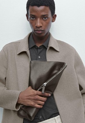 Pochette en cuir brun avec fermeture éclair, tenue par une personne portant un manteau en laine brun clair par-dessus une chemise sombre. Texture lisse et design minimaliste.