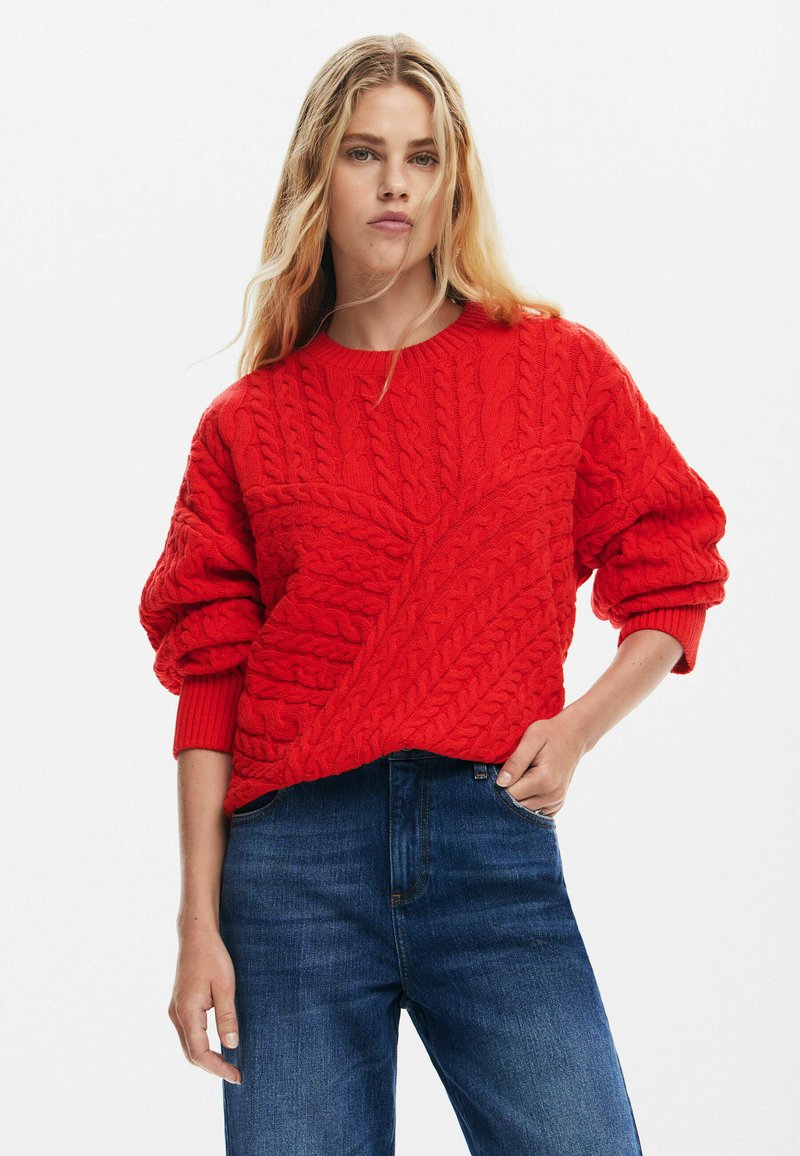 Maglione rosso lavorato a maglia con design a trecce, con spalle scese e polsini a coste, abbinato a jeans in denim blu scuro.