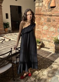 Ženska v črni obleki z enim ramenom z volančki in rdečimi čevlji stoji ob lesenem zunanjem mizi in stolih na kamniti terasi.